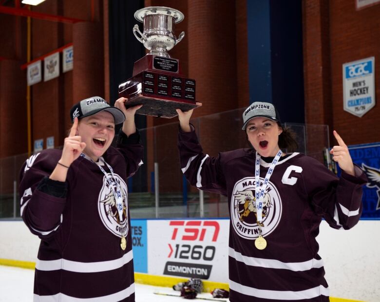 Two hockey players in their uniforms lift a trophy together and point their index finger up on the ice surface of a hockey arena.