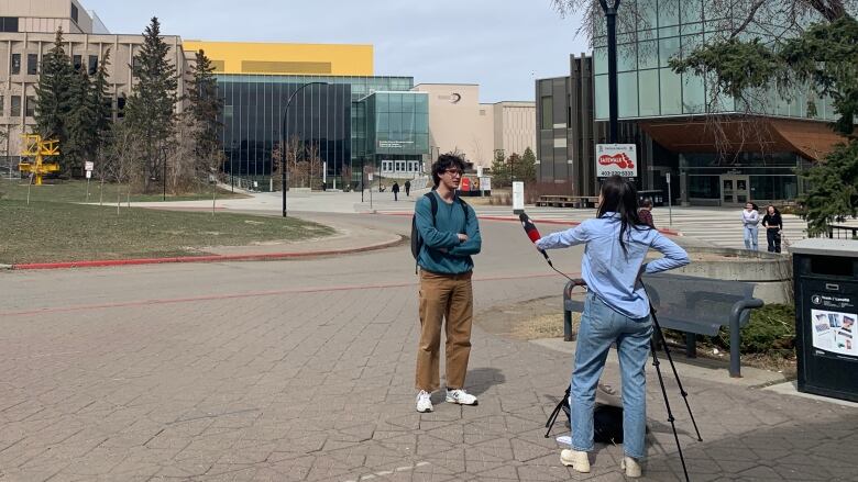 A woman with a camera interviews another man outdoors on a university campus.