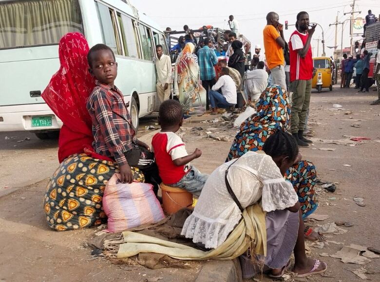 A group of children sit on a curb with luggage as people behind them board a van.