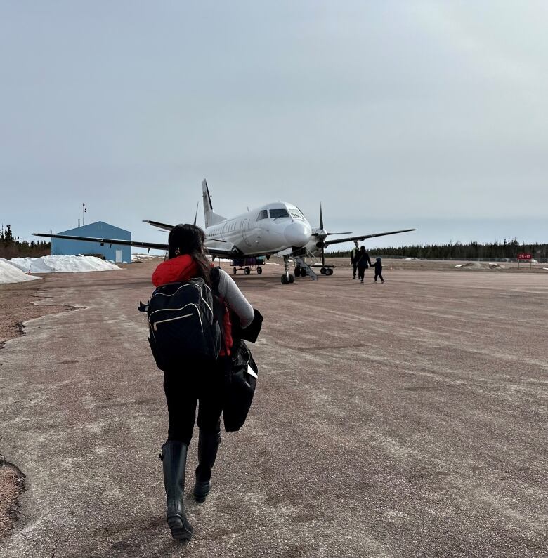 A woman is seen from behind, carrying a backpack, outside, walking toward a small airplane. 