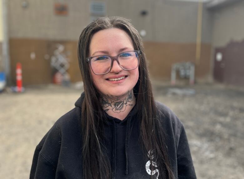 A person stands in a dirt lot, wearing glasses, long dark hear, and a floral neck tattoo.