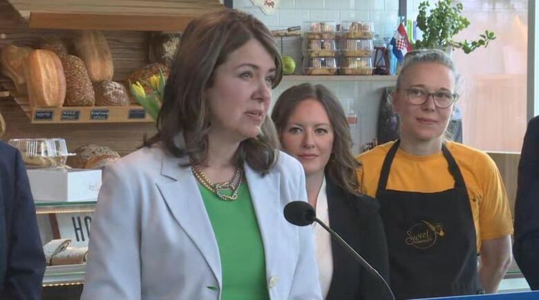 Danielle Smith stands before a microphone in a bakery wearing a green shirt and white jacket. 