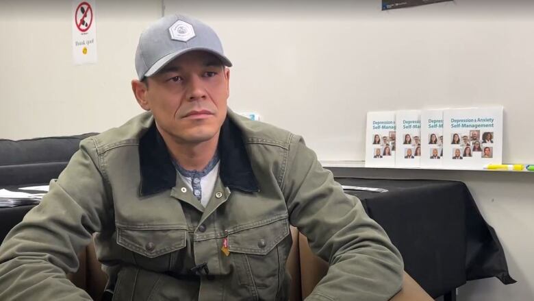 A man sitting with a denim jacket and an  Edmonton oilers baseball cap.