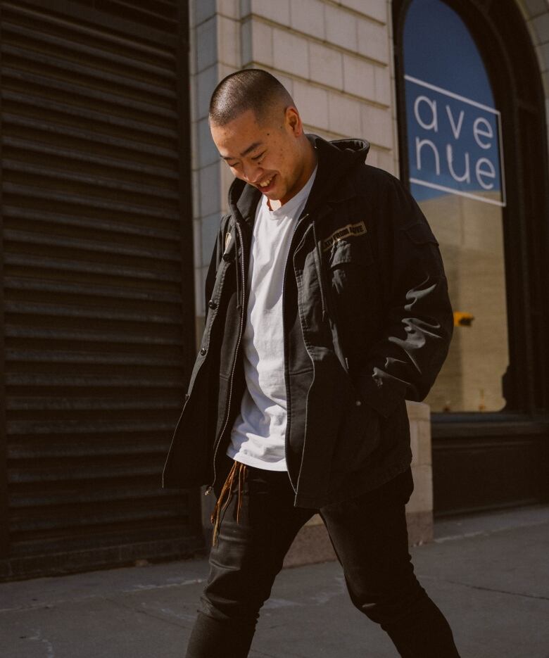 Set upon a building is a young man with his head down and smile on his face. He is wearing a white shirt beneath a green jacket.