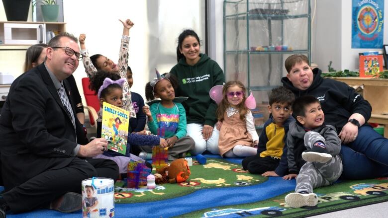 A man in a suit holds up a book and sticks his tongue out while posing for a photo with kids and early childhood educators.