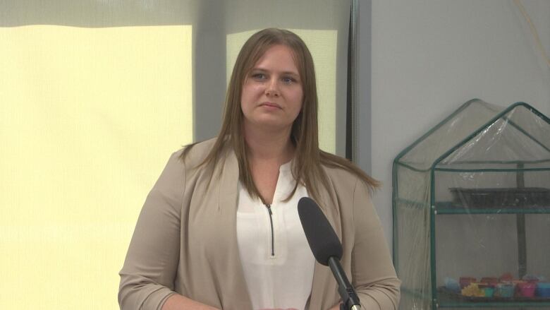 A woman in a white shirt and grey sweater speaks into a microphone at a childcare centre.