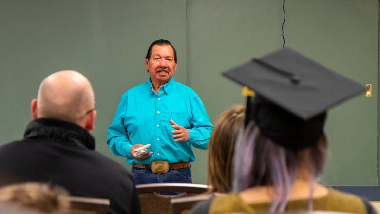 A man wearing a blue shirt speaks to a room of college graduates.