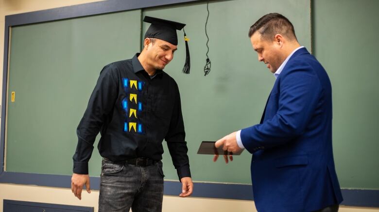 A man hands a graduate wearing a cap and ribbon shirt his diploma.