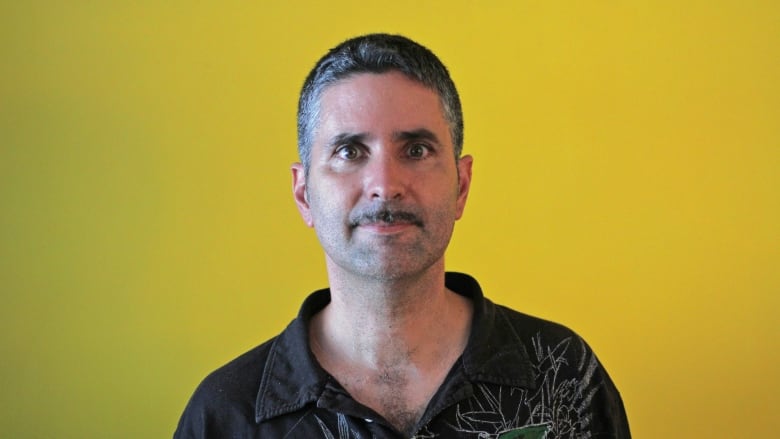 A man in a patterned black shirt poses for a photo against a bright yellow background.