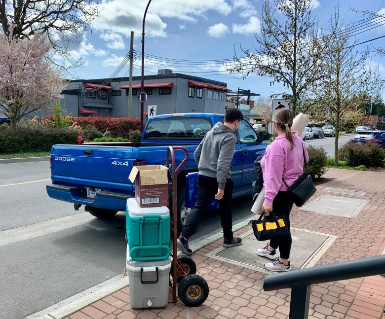 A man and a woman stand in front of a blue pickup truck. A dolly on the sidewalk is loaded with a box and two coolers. The woman holds a tool bag. 