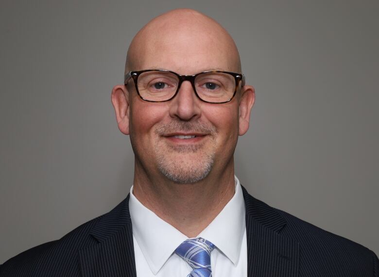 A man wearing a suit and black-framed glasses in front of a grey background