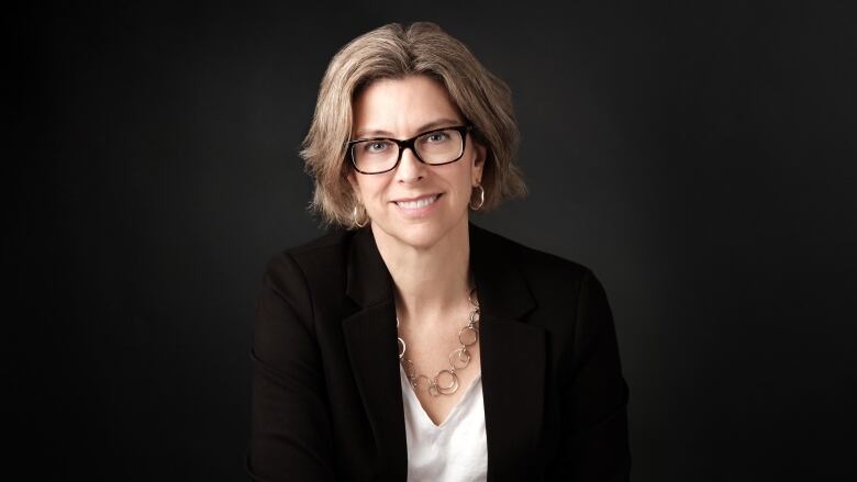 A professional headshot of a woman with short wavy blonde hair who is wearing black-framed glasses, a white blouse and black blazer. 