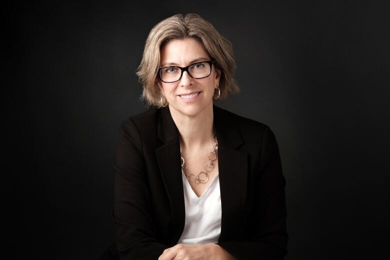 A professional headshot of a woman with short wavy blonde hair who is wearing black-framed glasses, a white blouse and black blazer. 