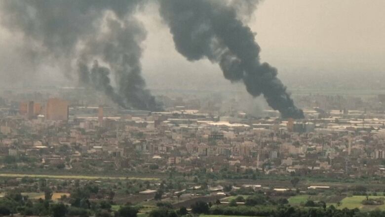 Two large black plumes of smoke rise above an urban vista.