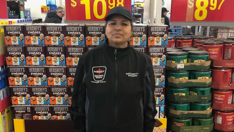 A woman wearing black pants, a black zip up jacket and a hat standing in the aisle of a store. 