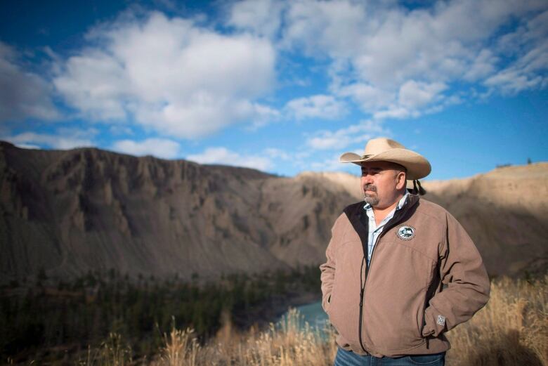 An Indigenous man wearing a cowboy hat poses in front of a hill.