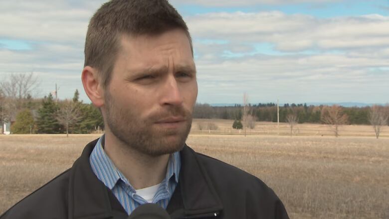 A man wearing a blue shirt and a black jacket stands in a field. 