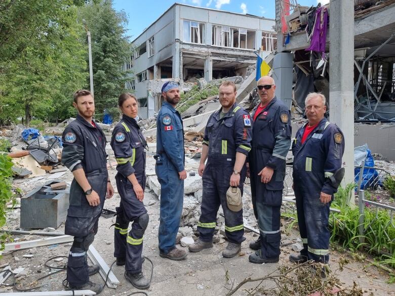 Six rescuers in uniforms pose in front of rubble from a building.