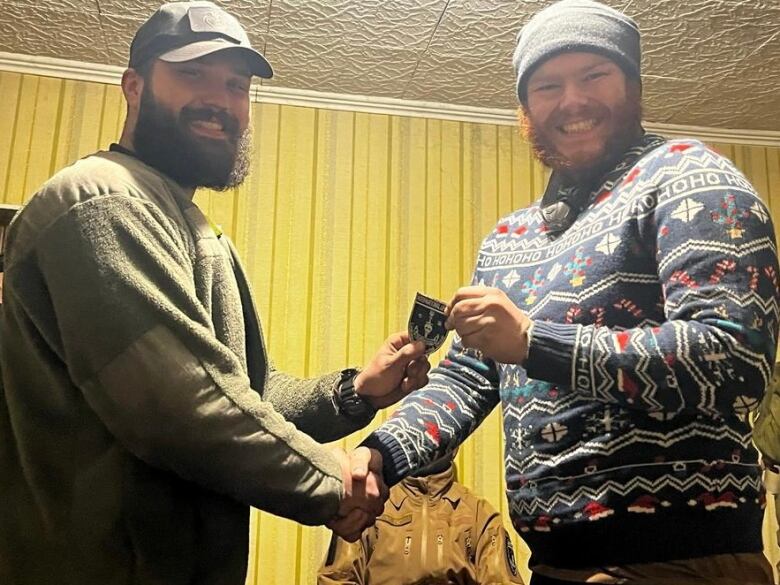 Two men wearing beards and hats smile and shake hands.