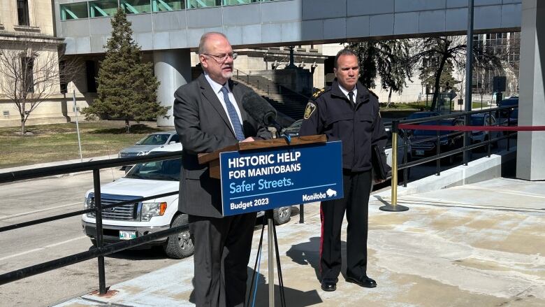 Two men, one in a grey suit at a podium, and one in a police uniform standing in the background, are seen outdoors in front of a skywalk.