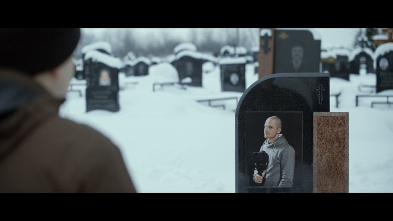 A man pictured from behind standing in a snow graveyard in front of a headstone with a photograph of a man holding a camera.