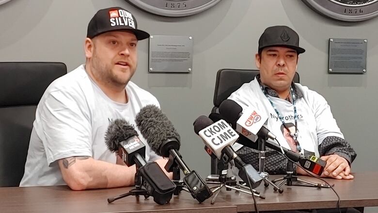 Two men wearing white t-shirts and black ballcaps are sitting at a desk, in front of multiple microphones.