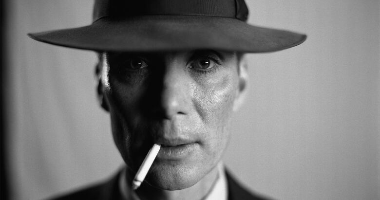 A black and white close up photo of a man looking directly into the camera while wearing a wide brimmed hat and smoking a cigarette. 