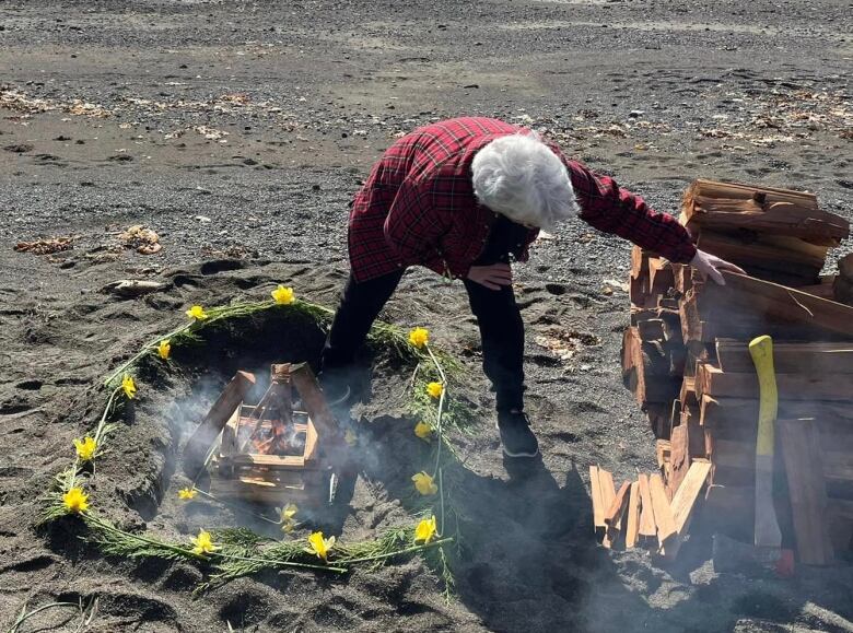 A woman tends to a fire on a beach that is surrounded by flowers and grass.