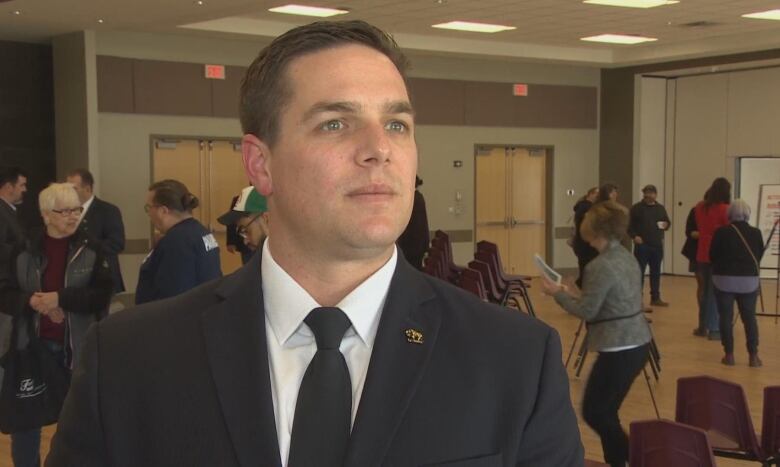 A man in a black tie, white collared shirt and black jacket speaks with media.