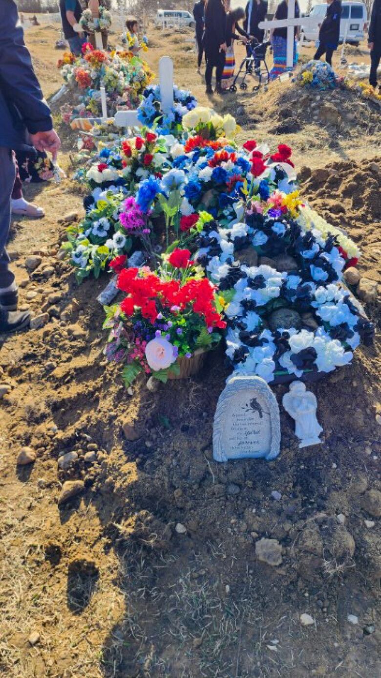 Bundles of flowers lay on top of a freshly covered grave. People are standing around the grave, paying respects.