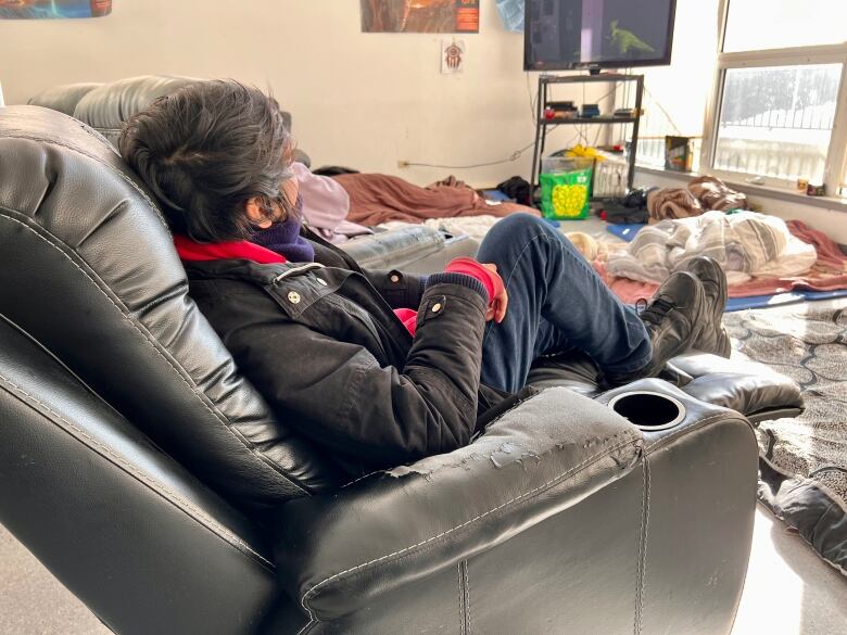 A man sits in a lounge chair with his back to the camera, wearing a black jacket and jeans with his feet up.