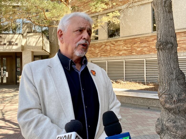 A man in a white suit and orenge shirt day pin speaks into microphones outside. 