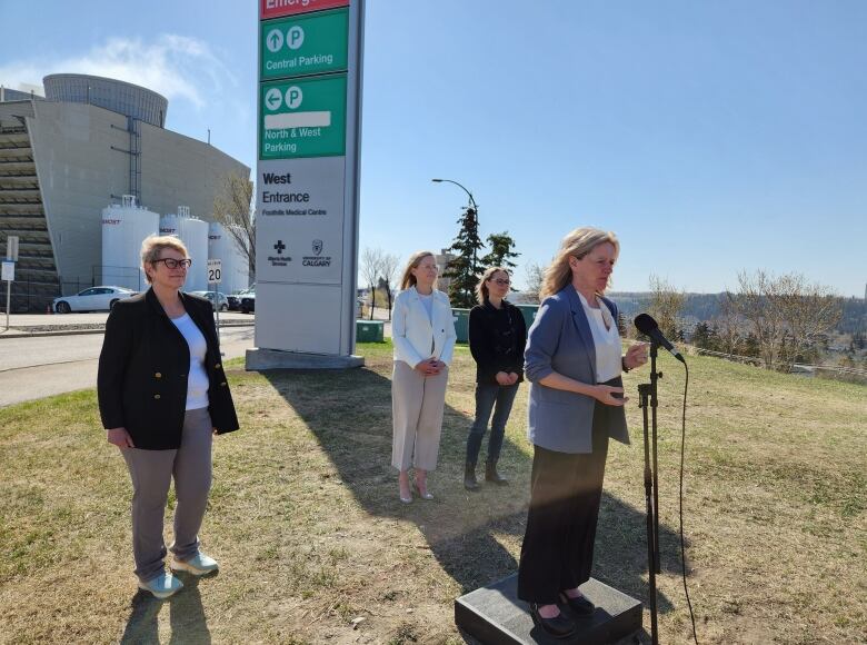 Rachel Notley speaks into a microphone in the shadow of a hospital sign, as three women who support her campaign look on.