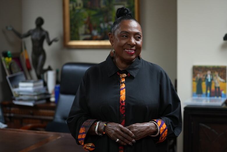 A woman wearing a black shirt with colourful trim clasps her hands and smiles as she stands in front of a desk in an personal office.