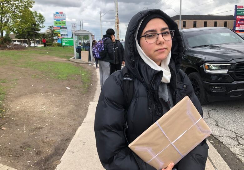 A girl wearing a white hijab and a grey sweater under a black jacket holding a brown package