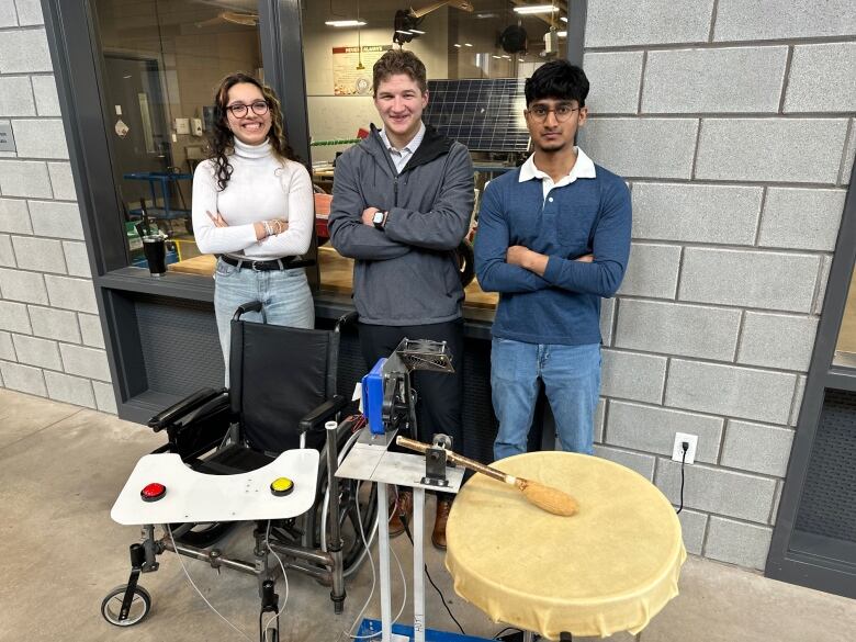 Three engineeering students stand in front of a wheelchair and a device that makes a stick hit a drum. 