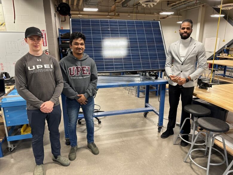 Three students stand in front of a solar panel in a big room at UPEI 