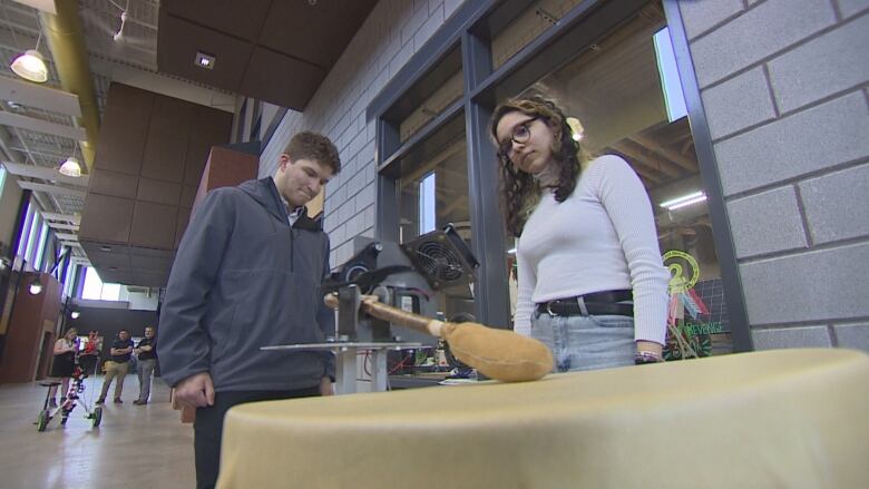 Two students look as a mechanized drumstick beats on a traditional drum. 