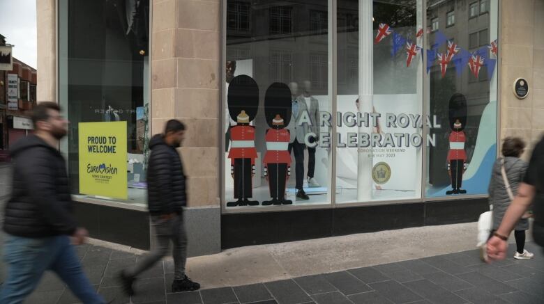 A corner window display of a business displays two signs, as pedestrians outside walk by. One is a yellow sign that reads: Proud to Welcome Eurovision. The other features the king's guards and the words: A Right Royal Celebration.