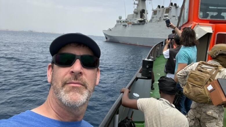 A man in sunglasses is seen on a tugboat as it nears a grey warship.