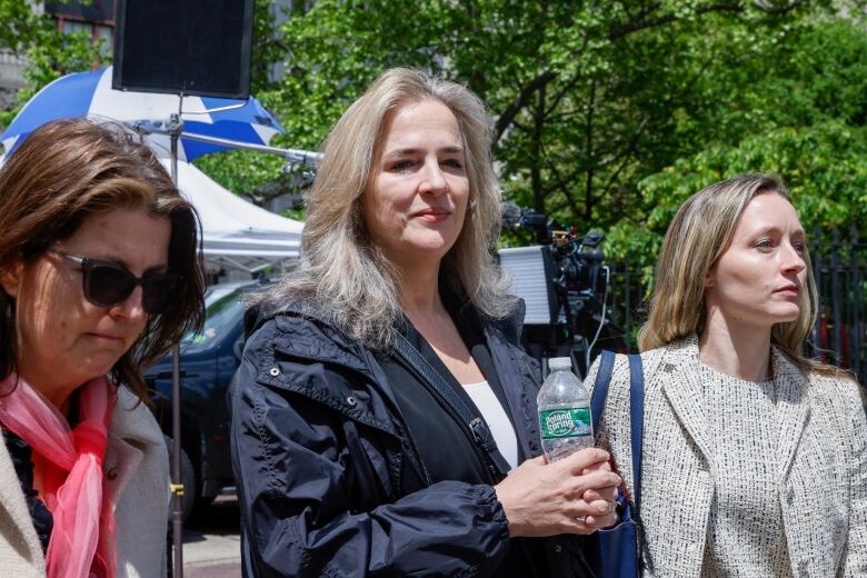 A woman with shoulder-length blond hair walks with 2 other women. 