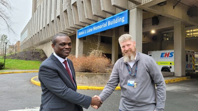 Two men shake hands in front of a concrete building underneath a blue sign that says 'Abbie J. Lane Memorial Building.' 