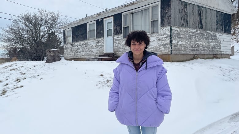 A young woman in a purple puffer jacket stands in front of a small bungalow with peeling paint in the wintertime.