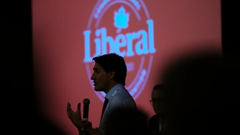 A man holding a microphone is seen in silhouette against a large screen that says 'Liberal.'