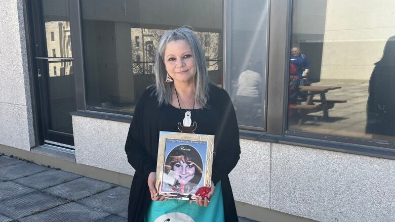 A woman with long grey hair wearing a colourful ribbon skirt looks at the camera as she holds a photo of a smiling younger woman.