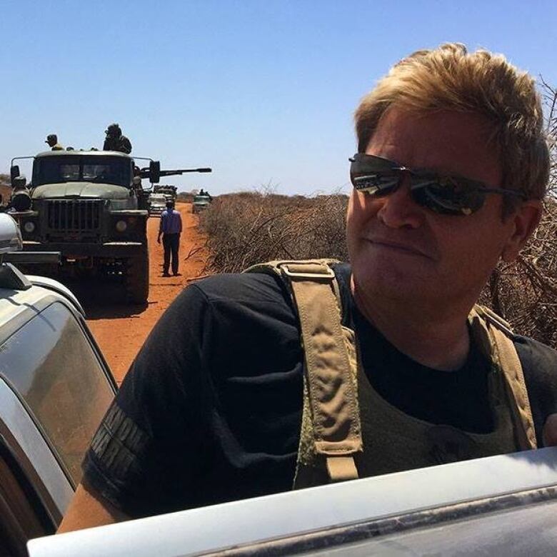 Man wearing a military vest and sunglasses stands outside a vehicle with soldiers and an armed tank and artillery in the background.