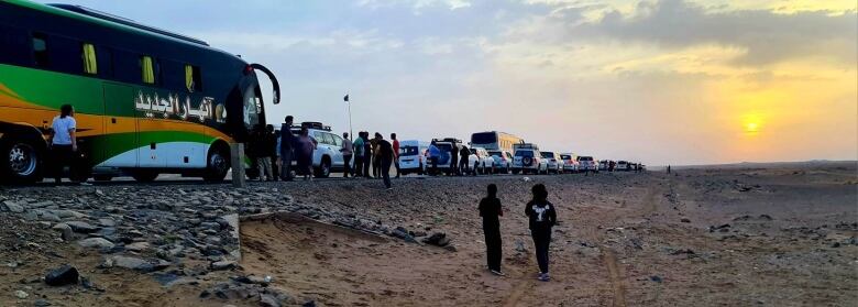 A long line of vehicles from buses to small cars line up on a desert road as the sun rises.