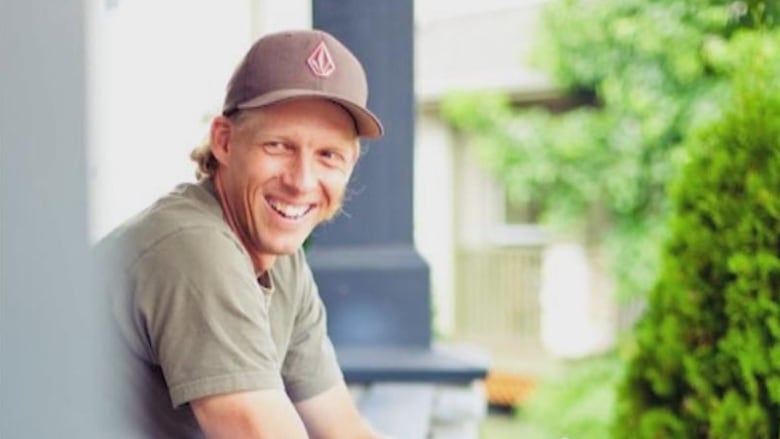 A man with orange hair, wearing a brown baseball hat, is leaning on his elbow over a porch. He is looking over his shoulder to the left and laughing. 