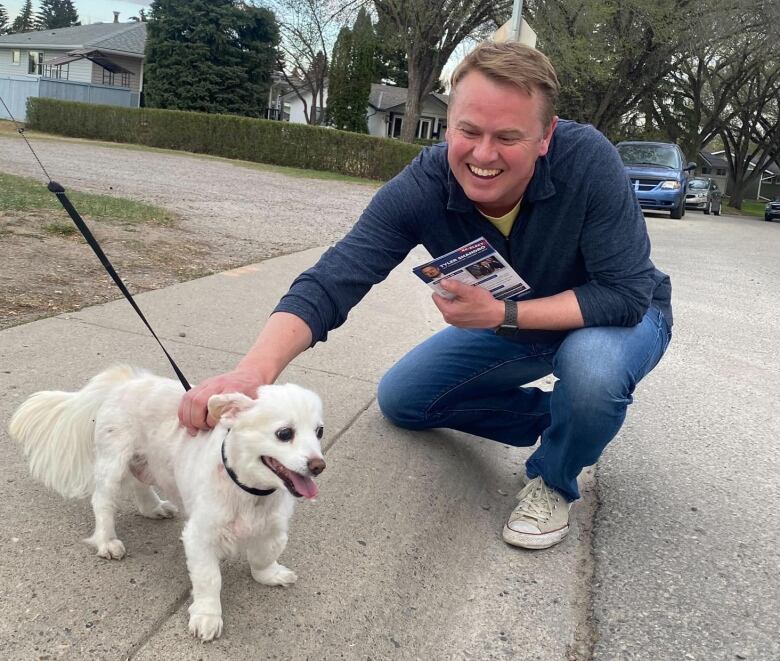 A man pets a little white dog on the sidewalk.