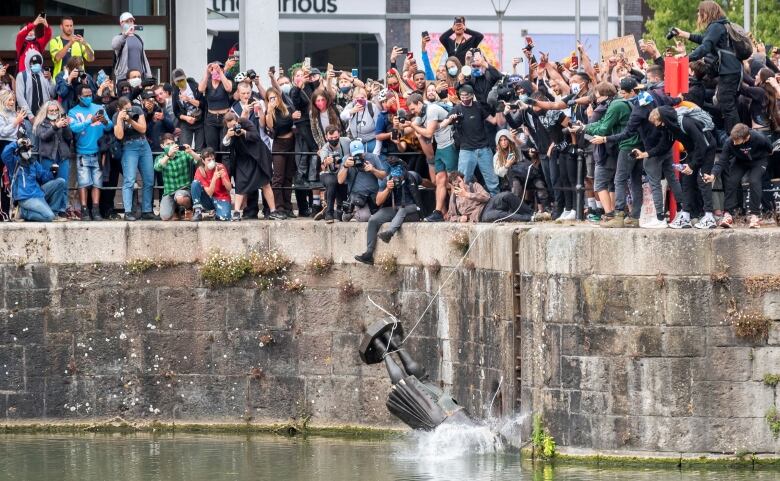 A large crowd of people cheers and takes photos as a statue falls into the water.